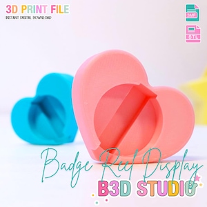Op de afbeelding: Een roze hartvormige badge reel display met een cirkelvormige opening en een blauwe hartvormige display op de achtergrond. De tekst "Badge Reel Display" en "B3D STUDIO" zijn zichtbaar. De tekst "3D PRINT FILE" is ook zichtbaar.