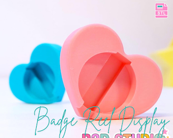 May include: A pink heart-shaped badge reel display with a circular opening and a blue heart-shaped display in the background. The text "Badge Reel Display" and "B3D STUDIO" are visible. The text "3D PRINT FILE" is also visible.