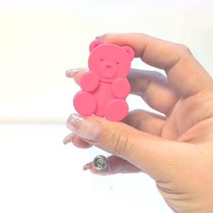 May include: A pink teddy bear-shaped badge reel. The bear has a simple design with rounded features and is attached to a clear plastic retractable reel with a metal clip. The background is white with a pink and white patterned border.
