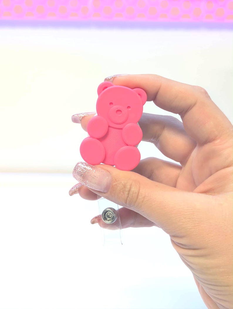 May include: A pink teddy bear-shaped badge reel. The bear has a simple design with rounded features and is attached to a clear plastic retractable reel with a metal clip. The background is white with a pink and white patterned border.