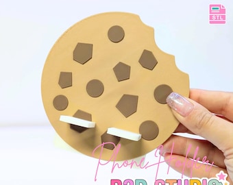 May include: A tan cookie-shaped phone holder with brown chocolate chip accents and a bite taken out of the side. The holder has two white support pieces. Text on the image reads "3D PRINT FILE" and "INSTANT DIGITAL DOWNLOAD".
