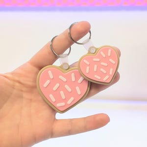 May include: Two heart-shaped keychains. The keychains are pink with white sprinkles and a tan border, resembling cookies. Each keychain has a silver ring attached to a clear plastic loop.
