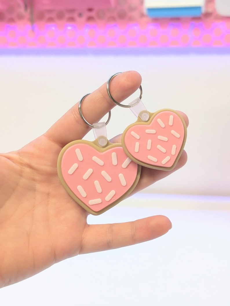 May include: Two heart-shaped keychains. The keychains are pink with white sprinkles and a tan border, resembling cookies. Each keychain has a silver ring attached to a clear plastic loop.