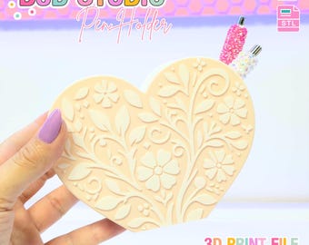 Puede incluir: Un portalápices en forma de corazón de color beige claro, con un diseño floral en relieve. El texto "B3D STUDIO Pen Holder" está en la parte superior. Dos bolígrafos son visibles en el portalápices. El texto "3D PRINT FILE" está en la parte inferior. Un archivo de impresión 3D.