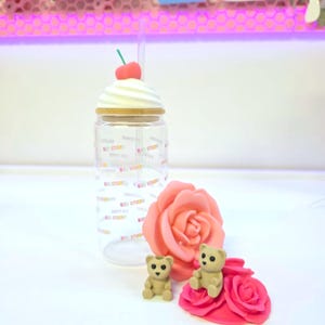May include: A clear tumbler with a whipped cream-shaped lid and a cherry on top. The tumbler has a straw and is decorated with the text "B30 STUDIO". Two small teddy bear figurines and a pink rose are also present.