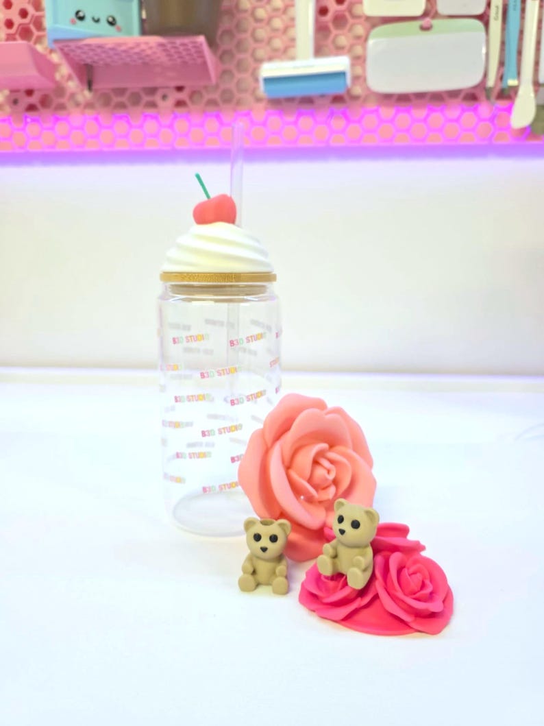 May include: A clear tumbler with a whipped cream-shaped lid and a cherry on top. The tumbler has a straw and is decorated with the text "B30 STUDIO". Two small teddy bear figurines and a pink rose are also present.