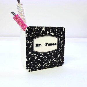 May include: A white pen holder with a black and white composition notebook design. The pen holder has the name "Mr. Funes" on the front. Two pens with crystal and pink rhinestone embellishments are inside the holder.