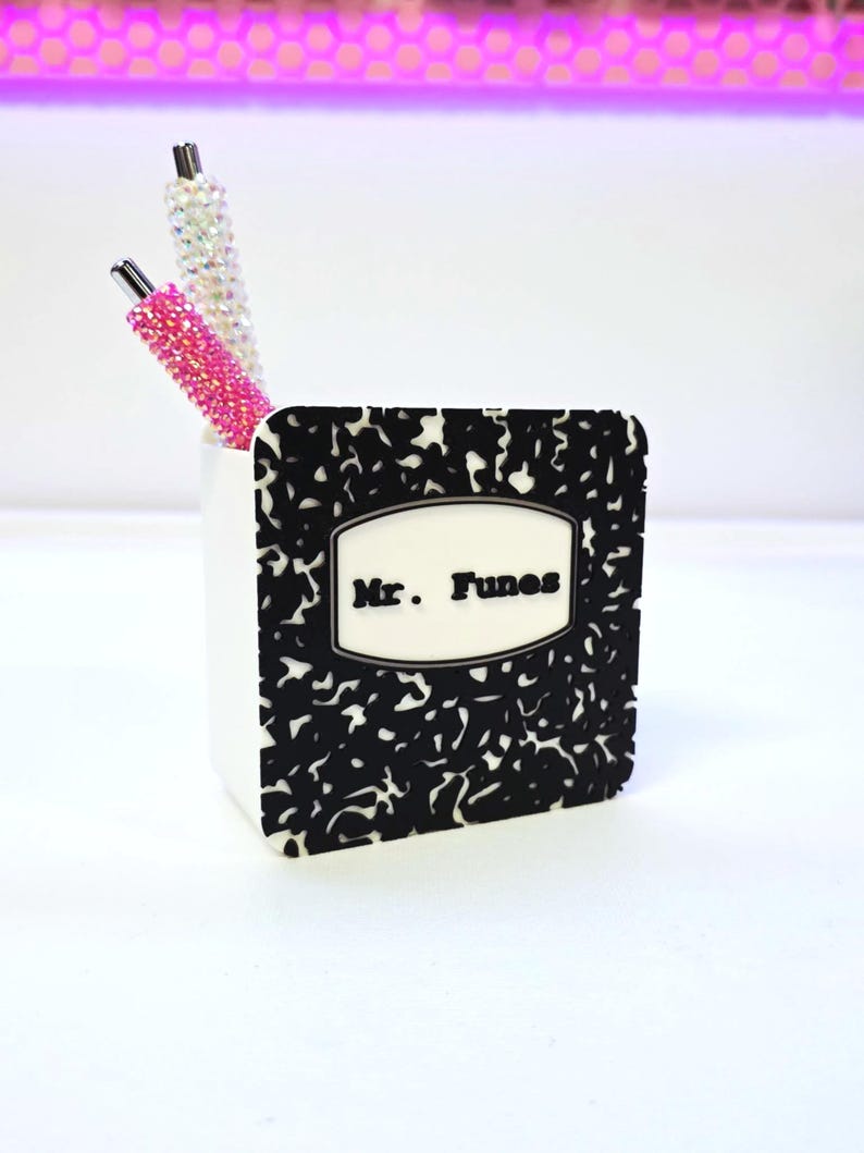 May include: A white pen holder with a black and white composition notebook design. The pen holder has the name "Mr. Funes" on the front. Two pens with crystal and pink rhinestone embellishments are inside the holder.