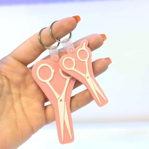 May include: Two pink scissor-shaped keychains with white scissor details. The keychains are attached to silver key rings. The background is a gradient of pink and white.