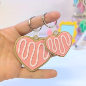 May include: Two heart-shaped keychains. The keychains have a pink heart design with a white squiggly line and a tan border. The keychains are attached to silver key rings with clear plastic holders.