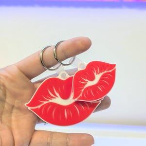 May include: Two red lip-shaped keyrings with white detailing. The keyrings are made of a smooth material and are held against a white backdrop. The design features a full pair of lips and a smaller single lip, attached to silver keyrings.