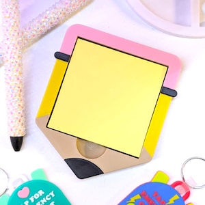 May include: A yellow notepad in a pencil-shaped holder with a pink border, yellow accents, and a brown base. Two pens with iridescent, crystal-like embellishments and two keychains with the text "TAP FOR EMERGENCY CONTACT" are also visible.