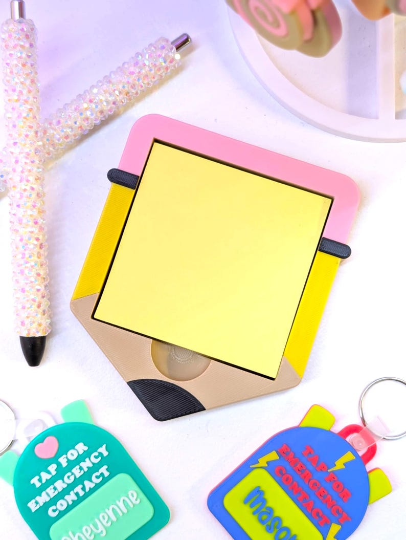 May include: A yellow notepad in a pencil-shaped holder with a pink border, yellow accents, and a brown base. Two pens with iridescent, crystal-like embellishments and two keychains with the text "TAP FOR EMERGENCY CONTACT" are also visible.