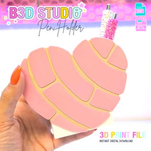 May include: A pink heart-shaped pen holder with a concha bread design. The pen holder has a light yellow outline and is held by a hand with orange nail polish. The text "B3D STUDIO Pen Holder" is at the top. The words "3D PRINT FILE" are at the bottom.