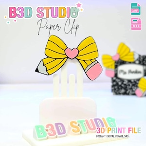 以下が含まれることがあります： ピンクのハートを中央に配置した、鉛筆のリボンの形をした3Dプリントのペーパークリップ。「B3D STUDIO Paper Clip」の文字が鉛筆リボンの上に表示されています。ペーパークリップのベースは白で、下部には「B3D STUDIO 3D PRINT FILE」の文字があります。