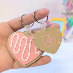 May include: Two heart-shaped keychains. One is pink with a white swirl design, the other is tan with a Spotify logo and star accents. Both have silver key rings and clear plastic attachments.