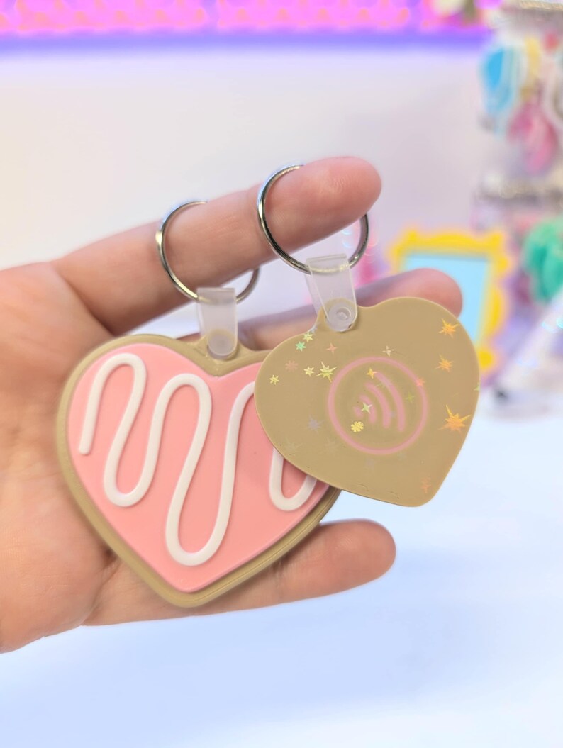 May include: Two heart-shaped keychains. One is pink with a white swirl design, the other is tan with a Spotify logo and star accents. Both have silver key rings and clear plastic attachments.