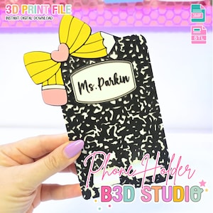 May include: A black and white phone holder designed as a composition notebook, with a yellow pencil and pink heart accent. The holder has the text "Ms. Parkin" and "Phone Holder B3D STUDIO" on it. The product is a 3D print file.