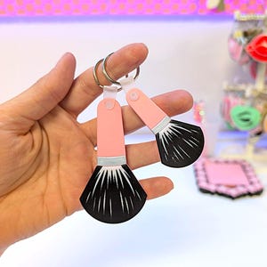 Può includere: Due portachiavi a forma di pennello da trucco. Ogni portachiavi presenta un manico rosa, una fascia argentata e una testa di pennello nera. I portachiavi sono attaccati a un anello argentato. Lo sfondo è sfocato, con accenni di rosa e bianco.