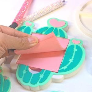 May include: A hand peels a pink sticky note from a cactus-shaped memo pad. The memo pad is teal and white with pink heart accents. Two pens with sparkly pink and white barrels are in the background. The items are on a white surface.