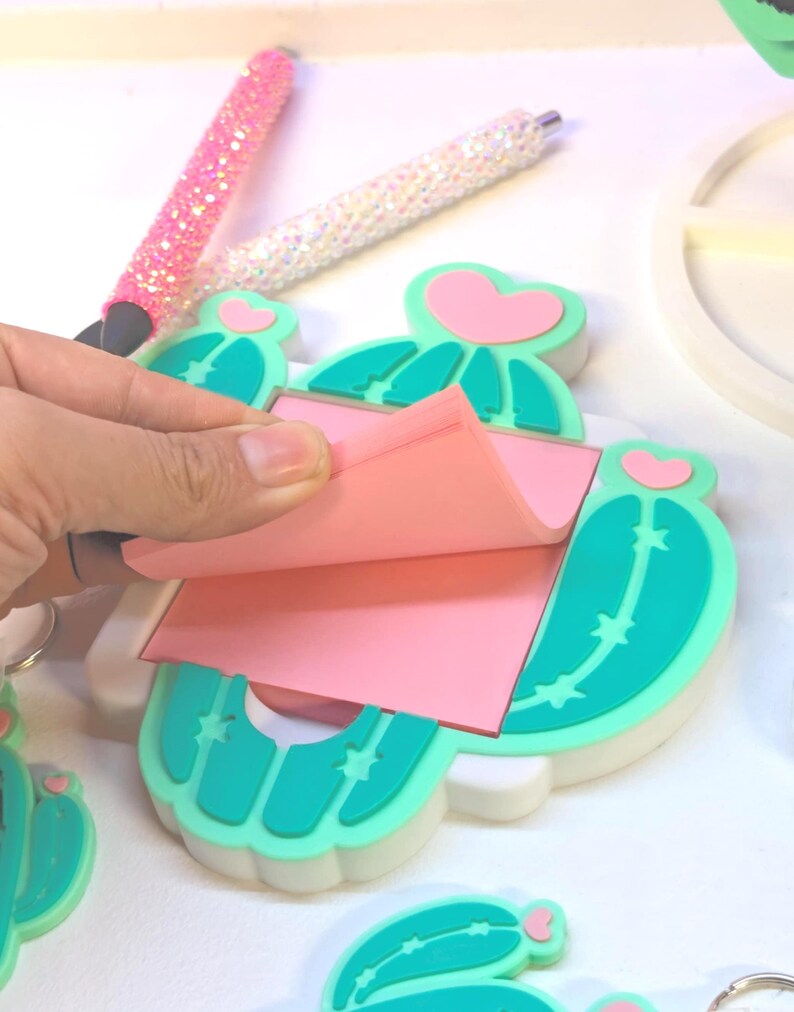 May include: A hand peels a pink sticky note from a cactus-shaped memo pad. The memo pad is teal and white with pink heart accents. Two pens with sparkly pink and white barrels are in the background. The items are on a white surface.