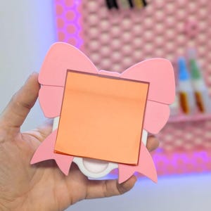May include: A pink bow-shaped memo holder with an orange square sticky note. The holder has a white base and is held in a hand. The background includes a pink wall with a polka dot pattern.