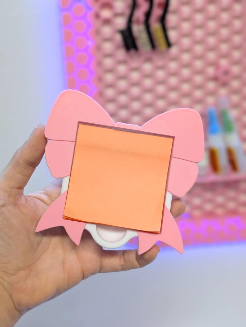 May include: A pink bow-shaped memo holder with an orange square sticky note. The holder has a white base and is held in a hand. The background includes a pink wall with a polka dot pattern.