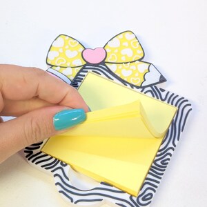 Peut inclure: Un porte-bloc-notes d&eacute;coratif avec un motif de n&oelig;ud papillon jaune et un c&oelig;ur rose au centre. Le support a une bordure z&eacute;br&eacute;e et contient une pile de notes autocollantes jaunes. Une main soul&egrave;ve une note.