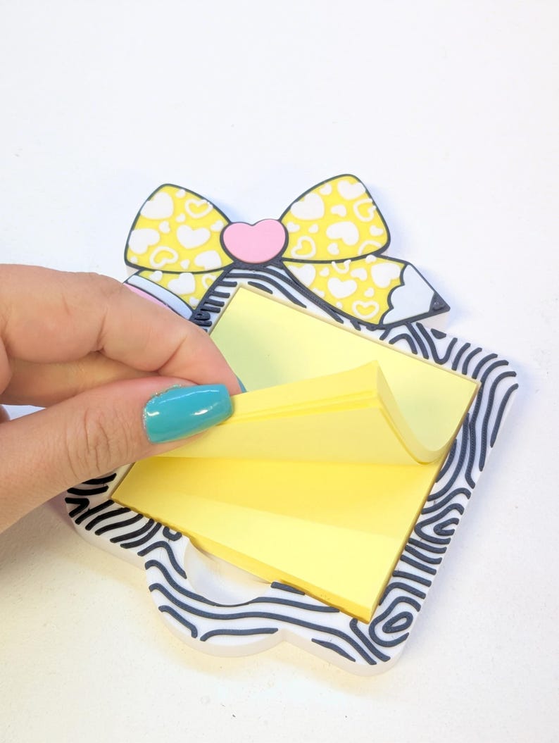 Peut inclure: Un porte-bloc-notes d&eacute;coratif avec un motif de n&oelig;ud papillon jaune et un c&oelig;ur rose au centre. Le support a une bordure z&eacute;br&eacute;e et contient une pile de notes autocollantes jaunes. Une main soul&egrave;ve une note.