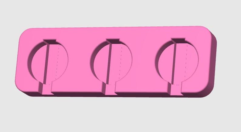 May include: A pink silicone mold for making three round-shaped treats. Each cavity has a vertical divider and a small stem at the bottom. The mold is rectangular with rounded corners and is designed for easy removal of the treats.