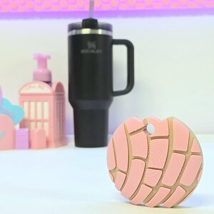 Puede incluir: Un posavasos rosa y beige con forma de pan dulce concha. Detr&aacute;s del posavasos hay un termo negro aislado con asa y pajita. Otros objetos decorativos rosas est&aacute;n en el fondo.