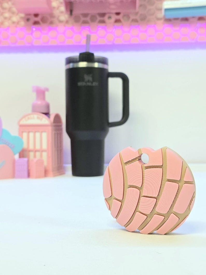 Puede incluir: Un posavasos rosa y beige con forma de pan dulce concha. Detr&aacute;s del posavasos hay un termo negro aislado con asa y pajita. Otros objetos decorativos rosas est&aacute;n en el fondo.