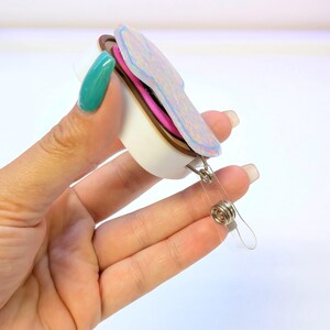May include: A retractable badge holder with a colorful, layered design. The badge reel features a white base, a brown middle layer, and a top layer with iridescent accents. A clear plastic strap and metal clip are attached.