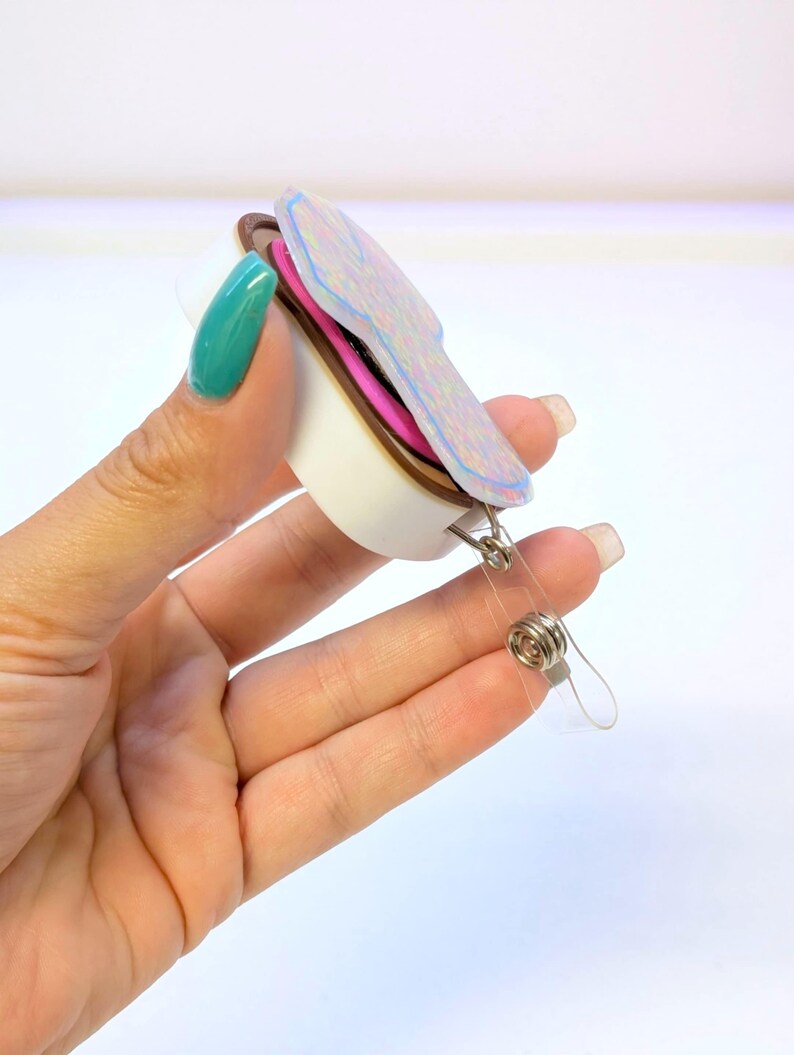 May include: A retractable badge holder with a colorful, layered design. The badge reel features a white base, a brown middle layer, and a top layer with iridescent accents. A clear plastic strap and metal clip are attached.