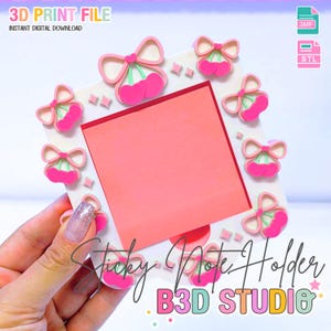 Op de afbeelding: Een vierkante sticky note houder met een roze notitie en kersen strik accenten. De houder is wit met roze en groene kersen en strikken langs de randen. Tekst op de houder luidt "Sticky Note Holder" en "B3D STUDIO".