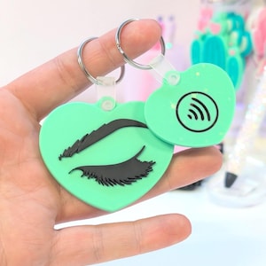 May include: Two heart-shaped keychains. One is mint green with a black eyelash design. The other is mint green with a black wireless symbol. Both have silver key rings and clear plastic attachments.