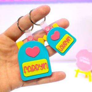 May include: Two blue backpack-shaped keyrings with pink hearts and yellow accents. One keyring reads "CARRYN" and the other reads "LAURYN" in yellow lettering. Both keyrings have silver key rings attached.
