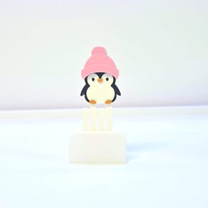May include: A small, white, rectangular object with three vertical prongs, topped by a cartoon penguin wearing a pink beanie. The penguin has a black body, white belly, and orange beak. The background is white.