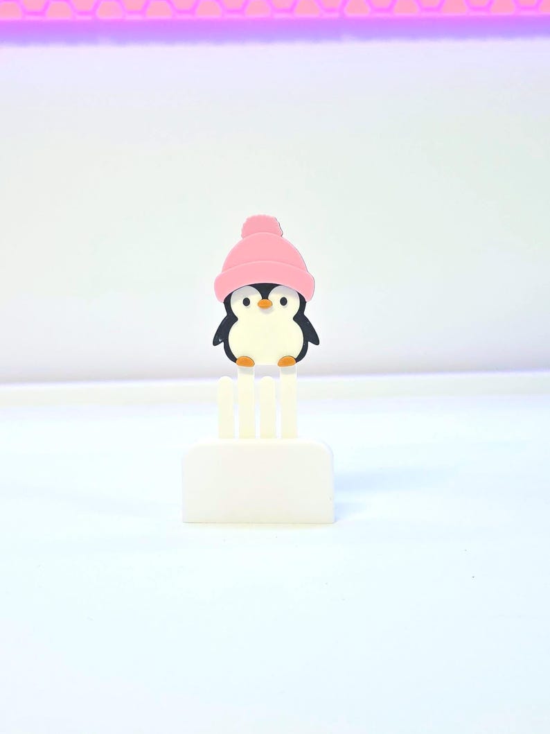 May include: A small, white, rectangular object with three vertical prongs, topped by a cartoon penguin wearing a pink beanie. The penguin has a black body, white belly, and orange beak. The background is white.