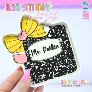 Puede incluir: Un joyero blanco con un diseño de cuaderno de composición en blanco y negro. El plato presenta un lápiz amarillo con una goma de borrar rosa y un corazón rosa. El texto "Ms. Parkin" se muestra en el cuaderno. También se ven las palabras "B3D STUDIO" y "Trinket Dish".