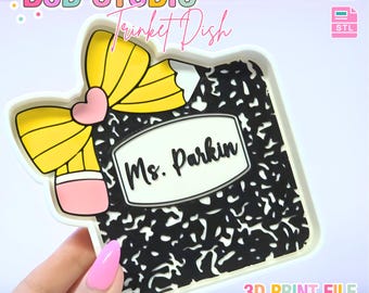 May include: A white trinket dish with a black and white composition notebook design. The dish features a yellow pencil with a pink eraser and a pink heart. The text "Ms. Parkin" is displayed on the notebook. The words "B3D STUDIO" and "Trinket Dish" are also visible.