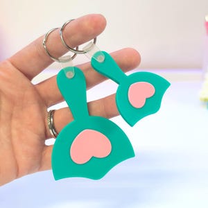 May include: Two teal keychains with pink heart accents. Each keychain features a rounded shape with a heart cutout and a small loop for the keyring. The keychains are held together by a silver keyring.