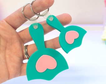 May include: Two teal keychains with pink heart accents. Each keychain features a rounded shape with a heart cutout and a small loop for the keyring. The keychains are held together by a silver keyring.