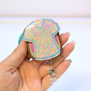 May include: A glittery badge reel featuring a pastel-colored shirt design with a blue outline. The badge reel has a brown and pink backing and a clear plastic retractable clip.