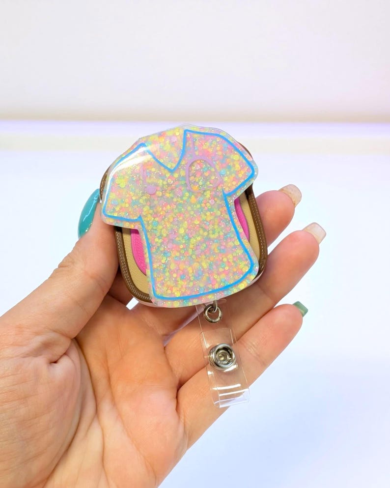 May include: A glittery badge reel featuring a pastel-colored shirt design with a blue outline. The badge reel has a brown and pink backing and a clear plastic retractable clip.
