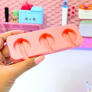 May include: A light pink silicone mold with three circular cavities, each with a central stem. The mold is held in a hand against a backdrop of pink and other colorful objects. The mold is designed for crafting or creating small, round items.