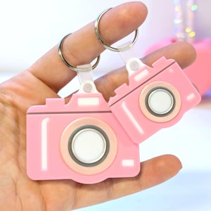Puede incluir: Dos llaveros rosas con forma de cámara con anillas plateadas. Los llaveros tienen un diseño de lente circular con un centro blanco y un borde rosa claro. Los llaveros se sostienen en una mano.