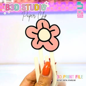 May include: A pink flower-shaped paper clip with a black outline and a white center. The paper clip is held by a hand with an orange painted nail. The text "B3D STUDIO Paper Clip" is at the top. The text "3D PRINT FILE" is at the bottom.