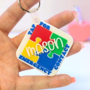 Pode incluir: Um porta-chaves branco com um design de peça de quebra-cabeça em vermelho, amarelo, verde e azul. O porta-chaves tem o nome "mason" em branco e as palavras "TAP FOR EMERGENCY CONTACT".
