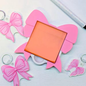 May include: A collection of pink bow-shaped keychains and a larger pink bow-shaped object with an orange square insert. A glittery pen rests nearby. The keychains and bow are on a white surface.